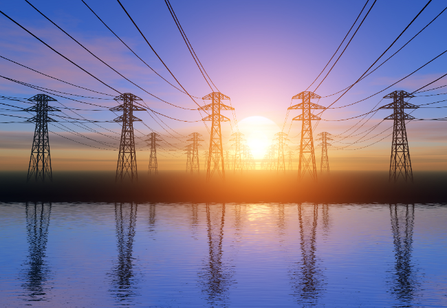 several electricity pylons, 5 in the foreground, with a sunset behind them and reflections in a lake beneath.