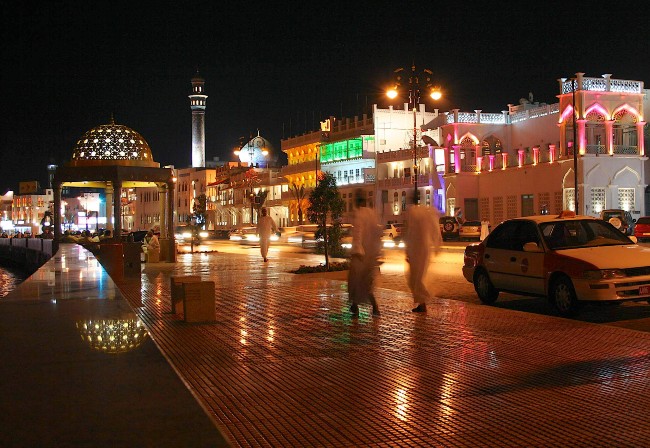 night shot of the main road alongside the harbour in Muscat. Several illuminated buildings and a mosque with traffic and passersby on the pavement.